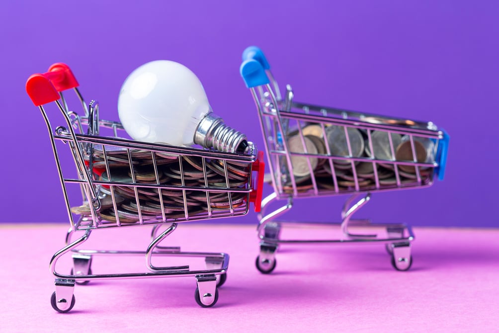 Two Trolley Carts Loaded with Currency Coins and Electric Lightbulb As Concept of Saving Energy. Over Purple Background.